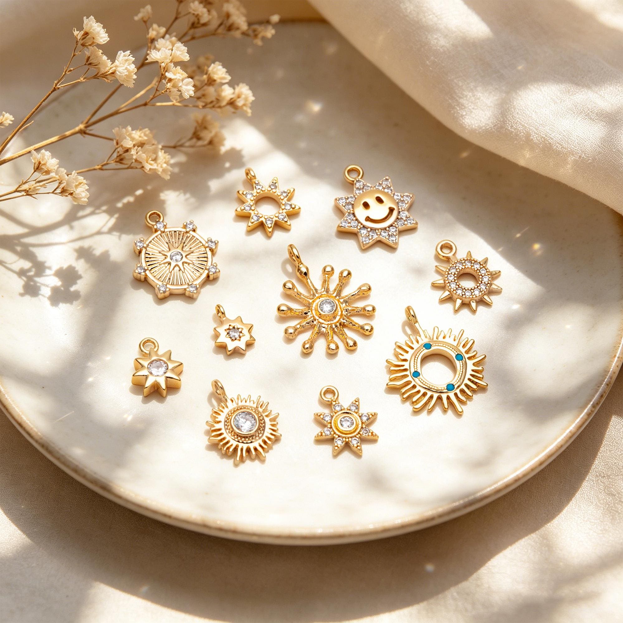 May include: A collection of gold-colored sun-shaped charms with various designs, including a smiling face, starburst patterns, and embedded crystals. The charms are displayed on a white plate, suggesting jewelry or craft supplies.